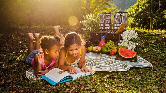 Two kids laying down to read a book on a blanket on the grass, while on a picnic